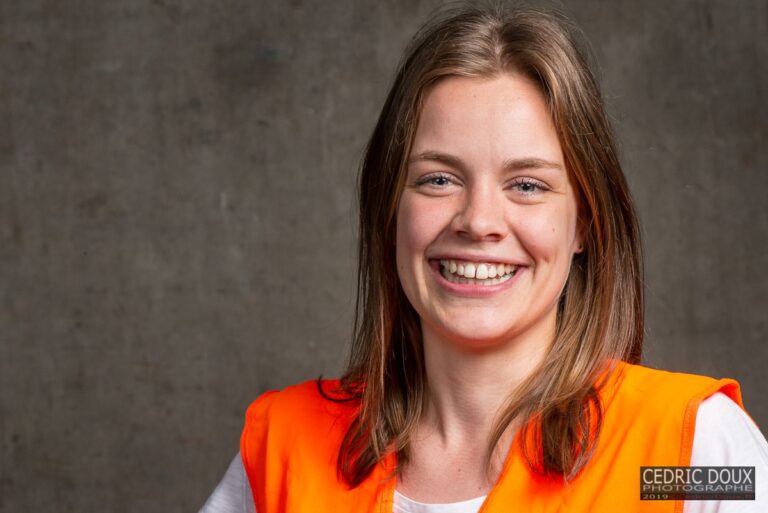 Jeune femme souriante en gilet orange haute visibilité devant un mur en béton d'entrepôt