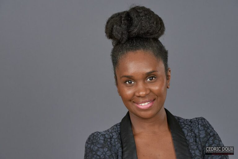 Femme aux cheveux noirs en chignon haut, veste à motifs et col noir, souriante sur fond gris