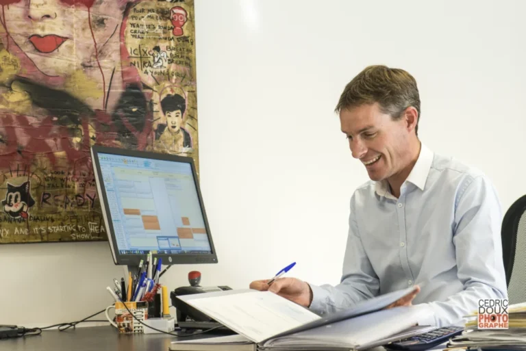 Homme en chemise bleu clair souriant et feuilletant un dossier à son bureau, écran et tableau d'art